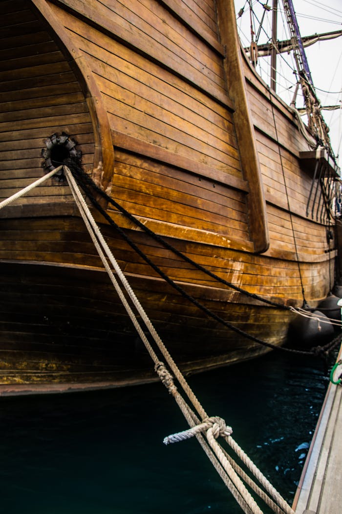 Home Close-up of an old wooden sailing ship docked at a port, showcasing ropes and nautical details.